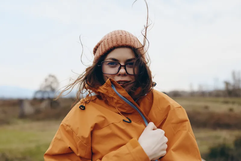 Woman lifestyle portrait outdoors, wearing orange jacket, glasses, and warm hat in windy countryside field, gentle smile, holding collar, soft daylight, travel mood fresh air, cloudy sky