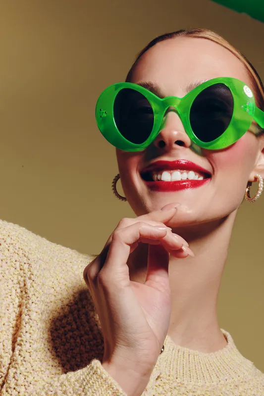 Smiling woman wearing large green sunglasses and red lipstick poses with hand near face. She wears a beige knitted sweater and gold hoop earrings on a neutral background. Fashion concept.