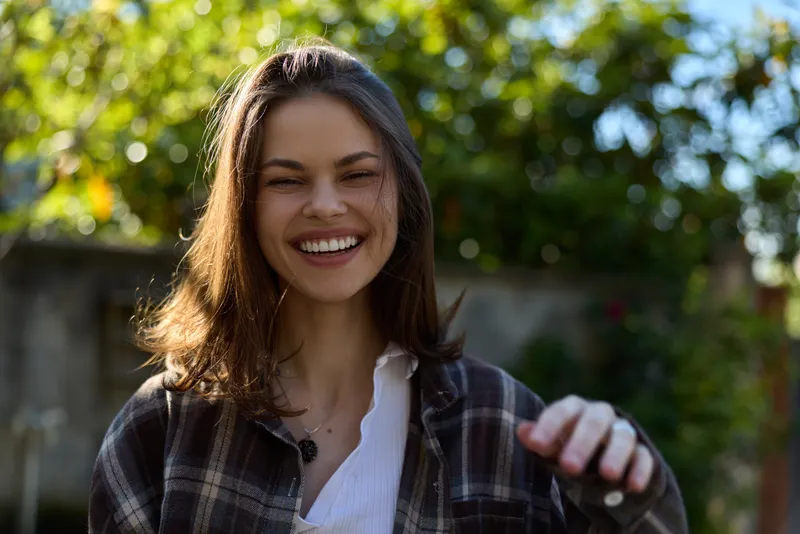 Cheerful woman outdoors, smiling with sunlit face, casual plaid shirt, relaxed posture, soft breeze, natural colors, lush greenery background in sunny garden setting, joyful