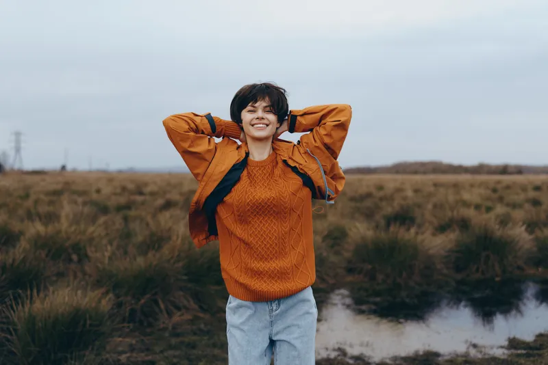 Smiling woman in an orange sweater and light jeans stands outdoors by a river in autumn fields, hands behind head, relaxed lifestyle portrait with natural wind and overcast sky