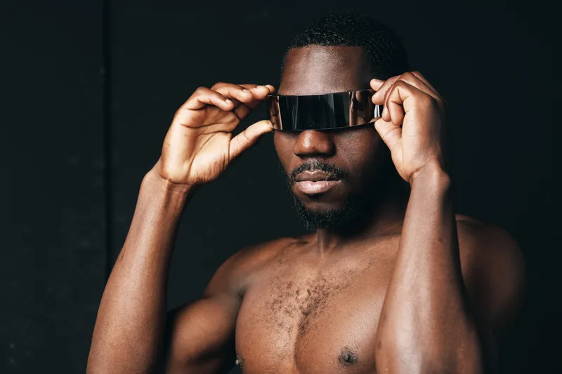 Black man with dark skin and beard wearing futuristic visor glasses in creative conceptual visual style. Portrait of confident African ethnicity male with bare chest on black background.