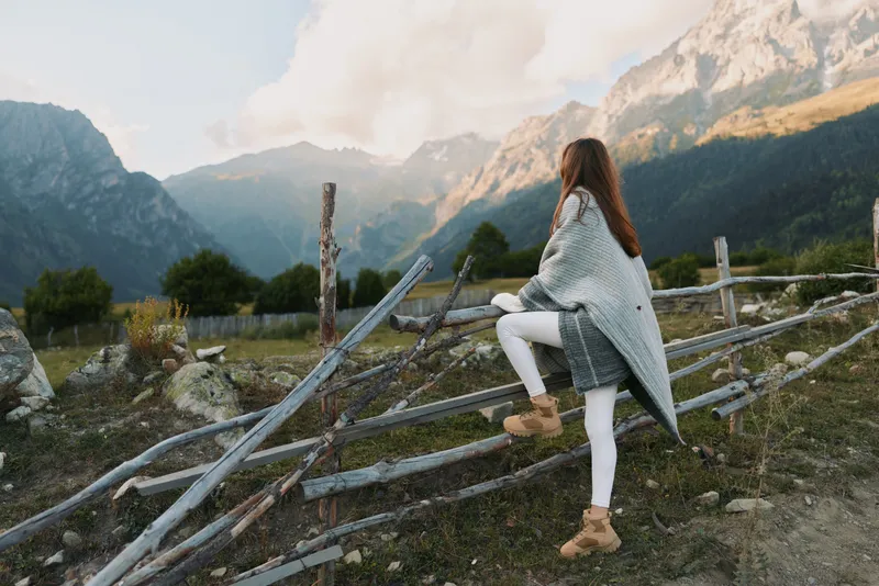 Young woman on a mountainside fence wearing a gray poncho and white pants, gazing into the distance with a calm, contemplative mood.
