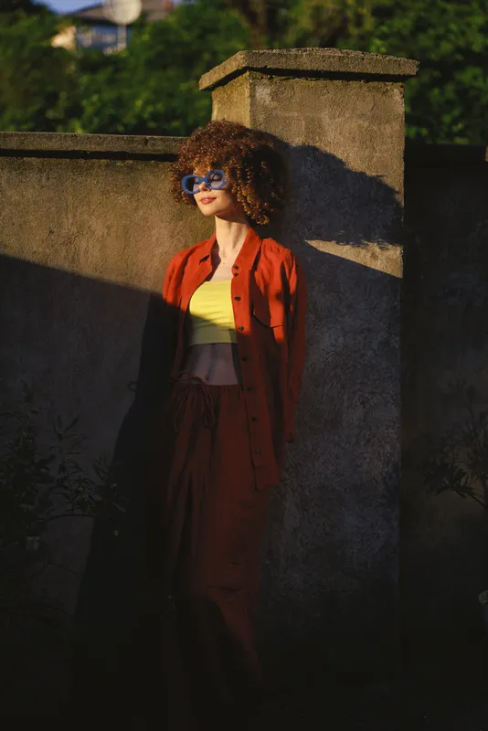 Smiling woman with curly hair wearing an orange jumpsuit over a yellow top, posing beside a stone wall outdoors. Casual lifestyle, fashion, and confidence