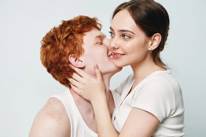 Couple, love, affection and young man kissing woman cheek with hands on face for romance and relationship with white background. Intimate, redhead and smile for happiness.