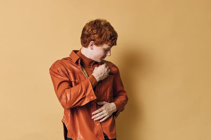 Young man with red hair wearing orange leather jacket standing against beige background looking sideways and adjusting clothing with hand on chest.