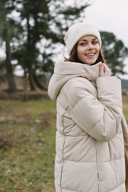 Winter woman in a beige puffer coat stands outdoors in a park, smiling, wearing a knit beanie, embracing cold weather with cozy style and soft natural light during late afternoon