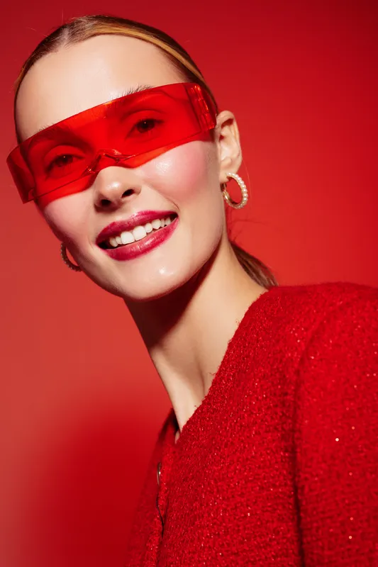 Smiling woman with red sunglasses and red outfit poses against red background. Fashion portrait with bright makeup and hoop earrings. Concept of style, confidence, and modern beauty.