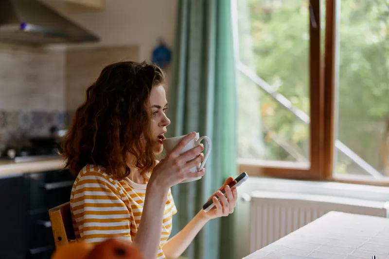 A woman sits indoors at home, sipping from a mug while glancing at her smartphone by a sunlit window, conveying a casual lifestyle moment of relaxed daily routine.