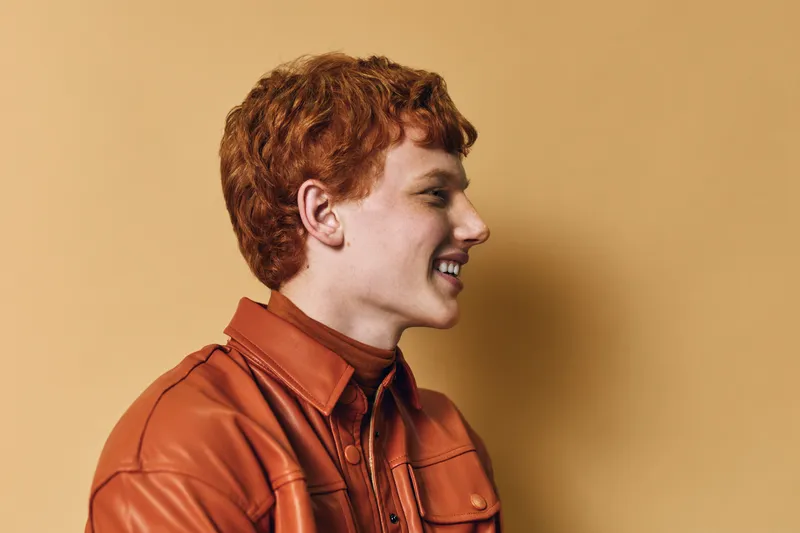 Redhair young man in orange leather jacket smiling showing profile on yellow background. Portrait of happy casual male model with curly hair and bright expression wearing stylish outfit.