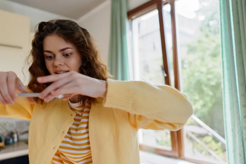Smiling woman with wavy hair reaching toward a surface at home, wearing a yellow cardigan and striped shirt, casual lifestyle scene, warm natural light, cozy domestic moment.