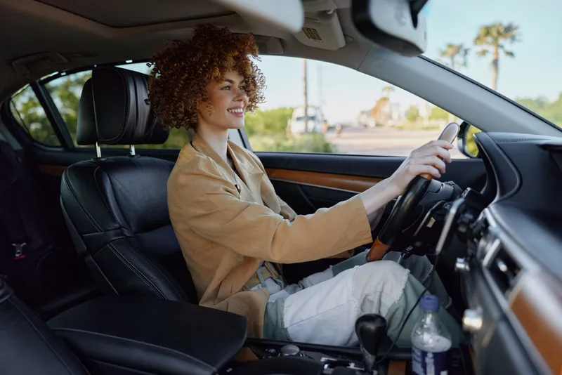 Young woman with curly hair sits behind the wheel of a car smiling softly as sunlight filters through the windows, creating a natural casual driving moment, a candid everyday scene.