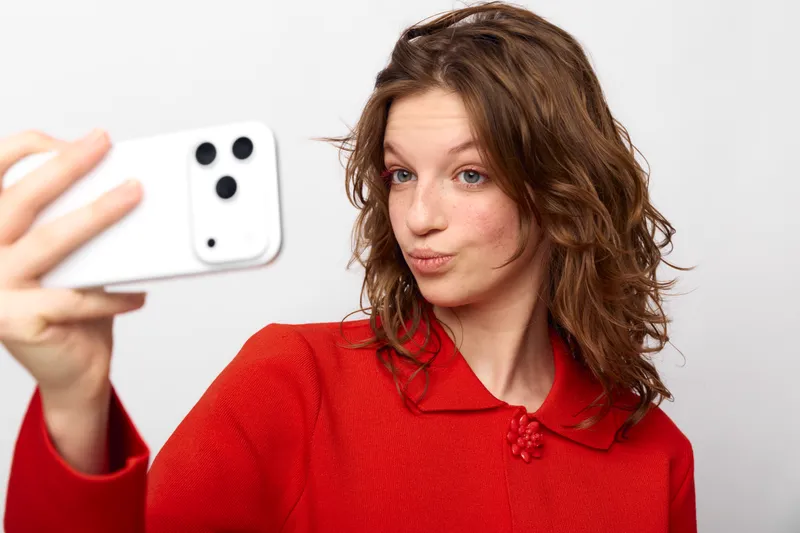 Young woman with curly brown hair wearing red sweater takes selfie with white smartphone on white background. Casual style, social media concept, modern technology and communication.