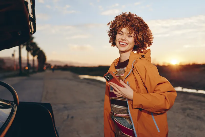 Cheerful woman outdoors lifestyle, laughing in orange jacket, curly hair glowing in autumn sunset, standing by beach shoreline, wind breeze, relaxed posture, warm natural colors