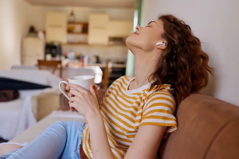 A woman lounges at home in a striped tee, sipping from a mug with wireless headphones, enjoying quiet moments. The scene conveys relaxed lifestyle, cozy comfort, and intimate home atmosphere.