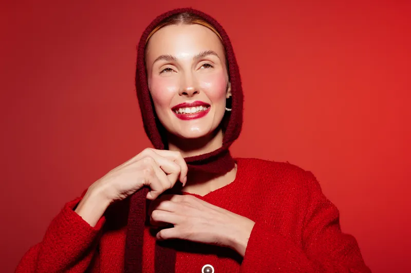 Smiling woman in red sweater and hood adjusts scarf on red background. Bright studio lighting highlights joyful expression and cozy winter fashion concept with warm colors.