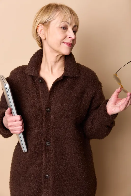 Senior woman in brown fleece holds smartphone and glasses, looks worried, seated indoors on plain background, conveys loneliness and communication stress, today