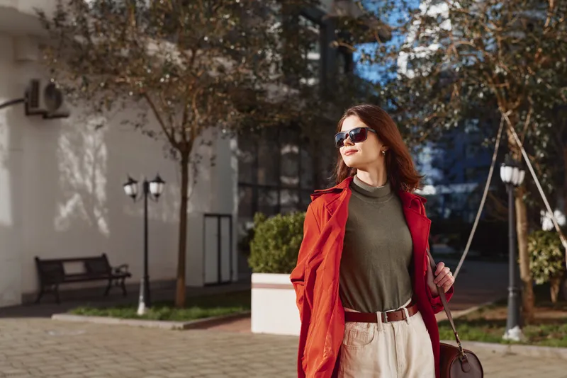 Stylish woman in a red coat and green top strolls confidently through a sunlit courtyard, wearing sunglasses and carrying a bag as autumn light fills the scene.
