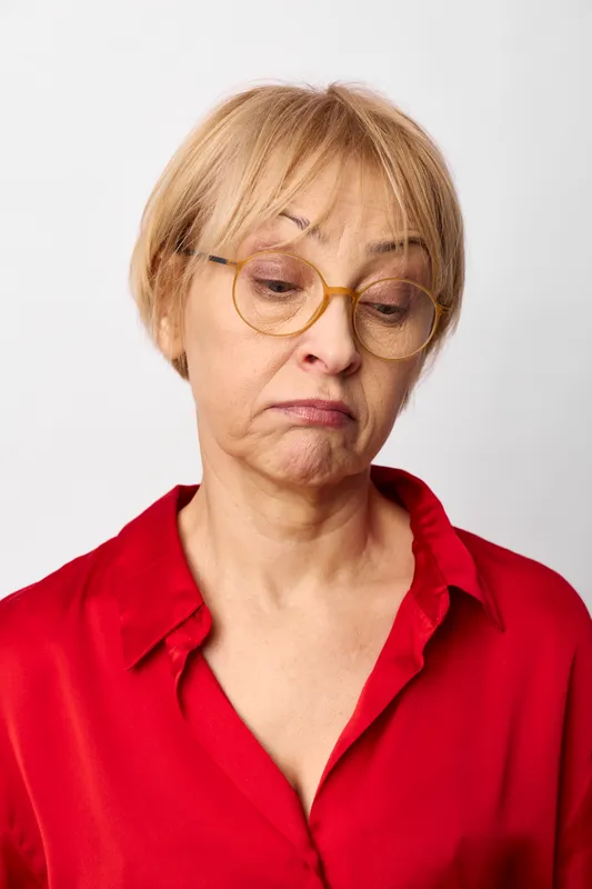 Sad elderly woman with glasses, studio portrait, sadness and emotion, white background, red shirt, short blond hair, wrinkles, closeup of tired stress, worried concern, aging