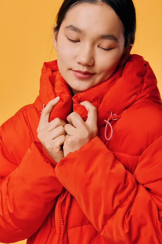 Young woman with closed eyes wearing bright orange puffer jacket on yellow background. Cozy winter clothing, warmth, comfort and casual style with peaceful expression and soft lighting.