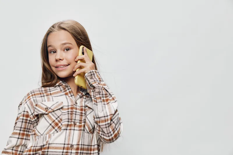 Girl with long hair smiling and talking on smartphone in plaid shirt on light background. Portrait of happy child using mobile phone for communication and casual conversation.