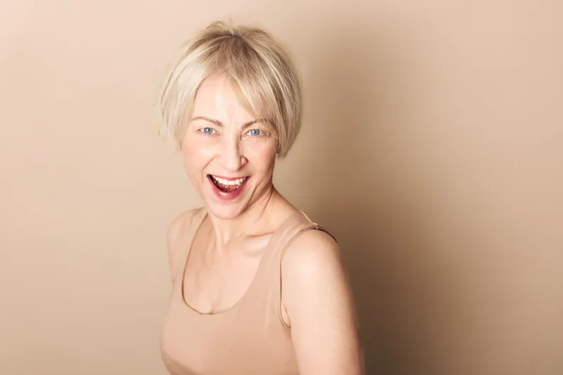 Happy middle aged woman with short blonde hair laughs joyfully wearing beige tank top against neutral background. Concept for positive emotions, confidence, and natural beauty in lifestyle.