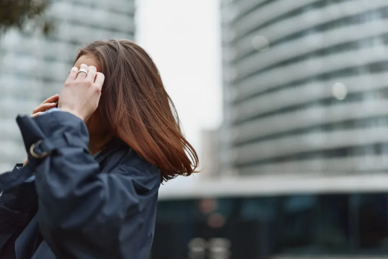 Young woman with brunette hair adjusts a navy jacket on a city street, the urban backdrop blurred behind her, conveying candid street style and focus.