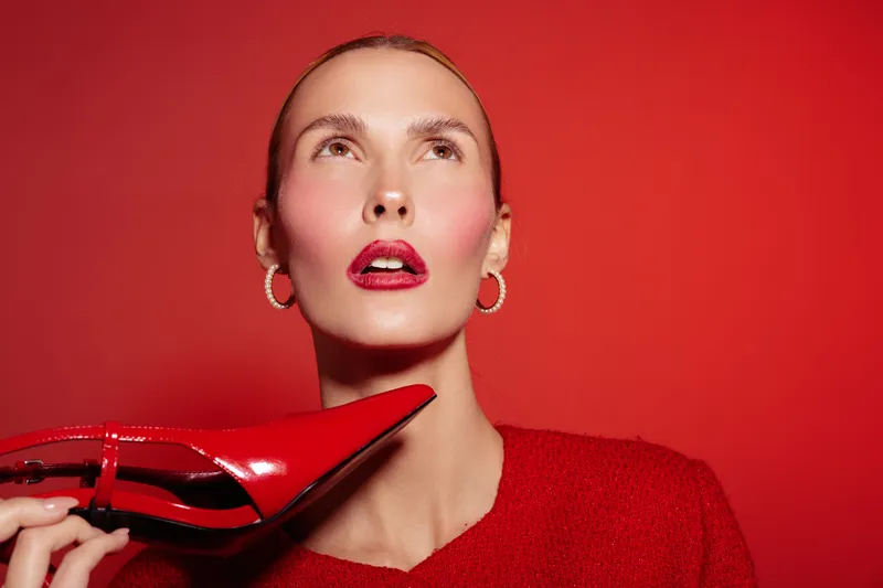 Stylish woman with red lipstick and earrings holds a red high heel shoe near her face against a red background. Concept of fashion, beauty, and bold style in studio lighting.