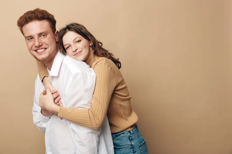 Smiling young couple embraces in casual clothes against beige background. The woman hugs the man from behind, both showing happy and affectionate expressions. Concept of love and togetherness.