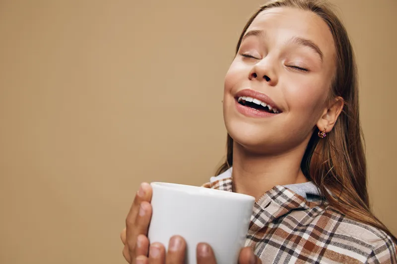Young woman with closed eyes enjoying hot beverage in white mug, cozy and relaxed with plaid shirt on beige background. Comfort, warmth and peaceful moment of happiness.