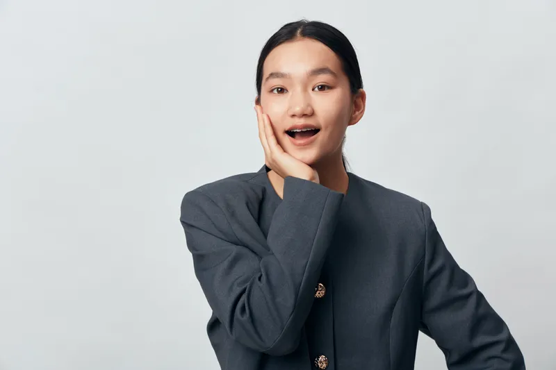 Young woman with dark hair in gray jacket expressing surprise with open mouth and hand on face against white background in studio portrait. Female person, casual style and emotion.