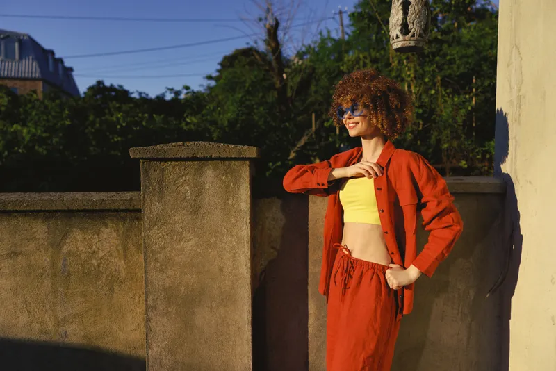 Smiling woman with curly hair wearing a bright yellow top and red jacket posing outdoor on a sunny street highlighting lifestyle, casual fashion, and confidence, portrait