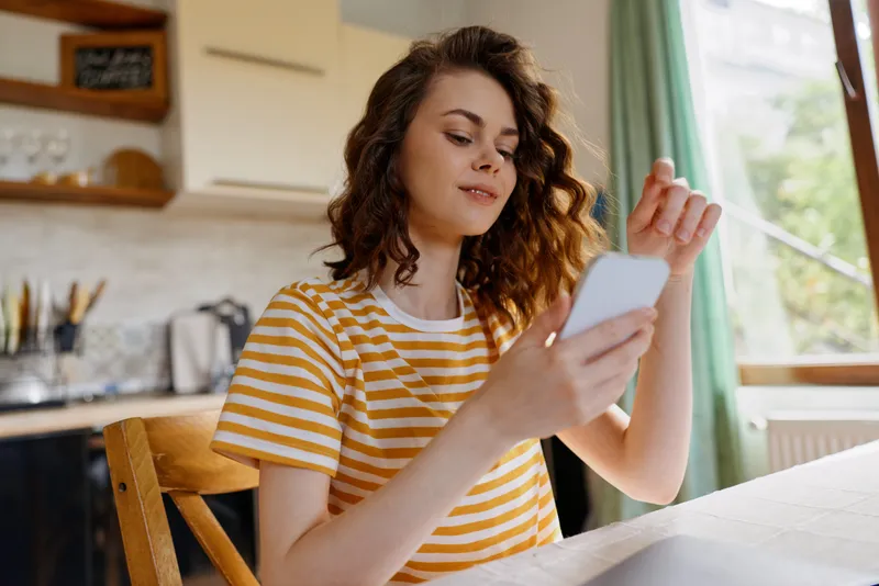 A woman wearing a striped yellow shirt sits at a kitchen table, looking at a smartphone. Natural light from a window creates a cozy home mood, highlighting casual lifestyle and daytime relaxation.
