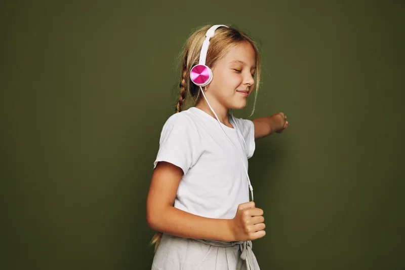Young girl wearing headphones enjoys music with closed eyes and relaxed expression on green background. Concept of happiness, relaxation, and childhood.