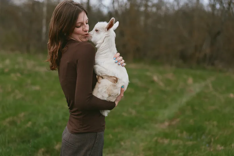 Goat kid held by a woman outdoors in a sunlit meadow, candid lifestyle moment, warm connection, soft breeze, natural colors, tranquil countryside background, gentle embrace, peaceful scene, authentic