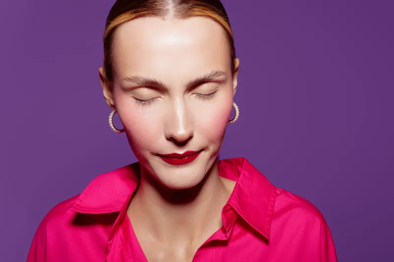 Close-up portrait of a woman with closed eyes wearing red lipstick and pink shirt against purple background. Concept of beauty, makeup, fashion, and calm expression.