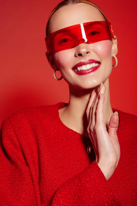 Smiling woman in red sweater and futuristic red sunglasses poses against red background. Fashion portrait with bright makeup and hoop earrings. Concept of style, confidence, and modern accessories.