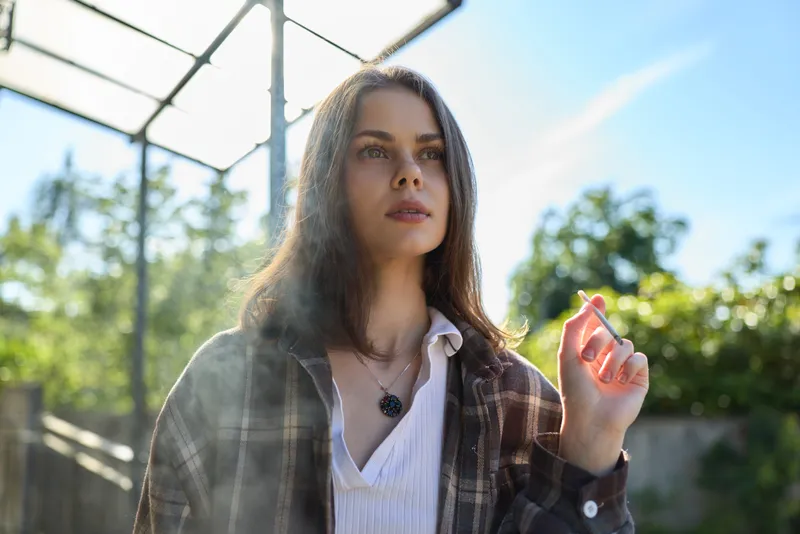 Brunette woman in a plaid jacket over a white blouse stands in a sunlit outdoor setting, looking upward with a thoughtful, calm expression, embodying casual fashion in natural light