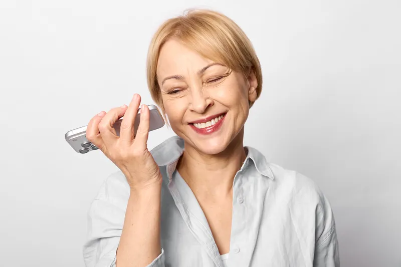 elderly woman smiling while holding phone to her ear, joyful call expression, clean studio background, communication and customer service concept, friendly support call