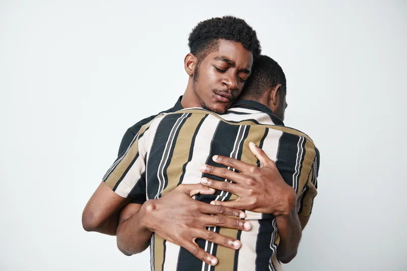 Two young black men with dark skin hugging each other in emotional support and friendship, wearing casual striped shirts, isolated on white background.