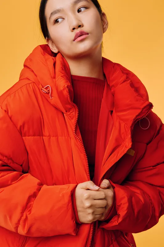 Young woman wearing bright orange winter jacket and sweater posing against yellow background in studio. Female model looking aside holding jacket zipper with both hands for fashion portrait.