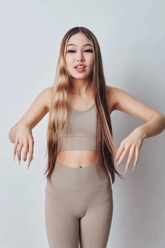 Young woman with long hair poses in beige sportswear outfit on neutral background for fitness, wellness and active lifestyle concept. Model with natural makeup shows confident expression.
