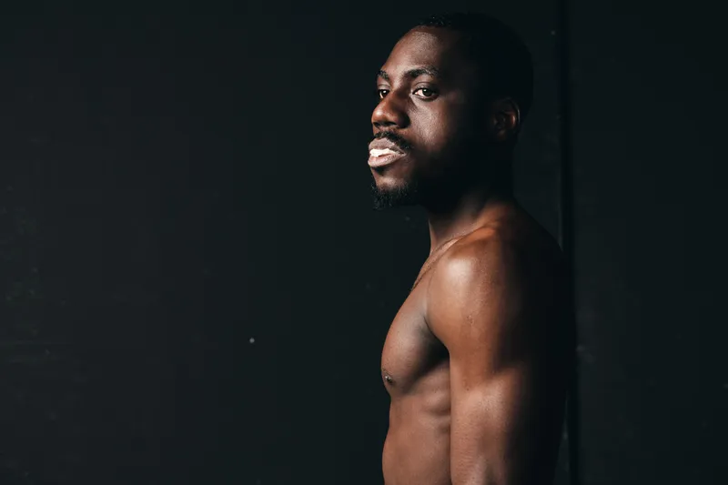 African man with dark skin and muscular body posing shirtless in creative conceptual visual style against black background. Portrait with dramatic lighting and serious expression.
