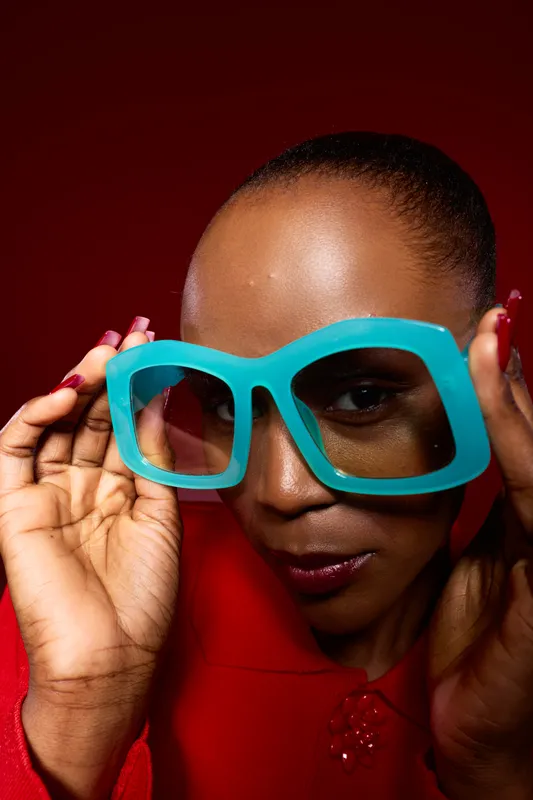 Close-up of an African woman with dark skin holding oversized blue glasses on a red background. Portrait of confident black female model with smooth skin and glossy lips in studio lighting.