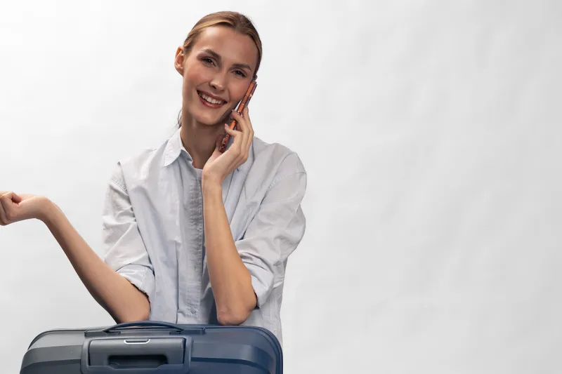 Smiling woman with suitcase talks on phone, wearing casual shirt. Travel concept with positive mood and communication. Isolated on white background with natural lighting.