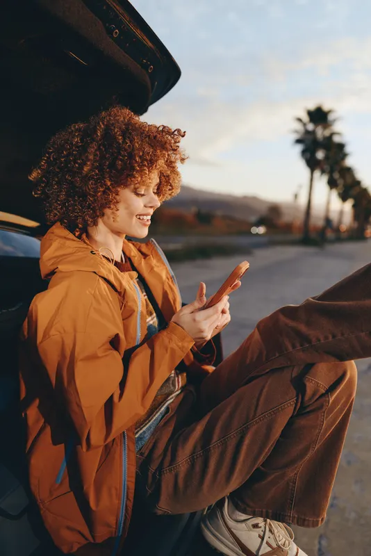 Woman lifestyle travel scene, curly hair woman in orange jacket by car open door, smiling and using smartphone, golden sunset light, road with palm trees, outdoor portrait, relaxed