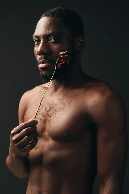 Creative conceptual portrait of African man with dark skin holding dried flower near face in studio. Artistic visual style with bare chest and thoughtful expression on black background.