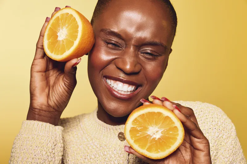 Happy African woman with dark skin holding two halves of a fresh orange near her face. Smiling female model in beige sweater on yellow background. Concept of health, wellness, and natural beauty.