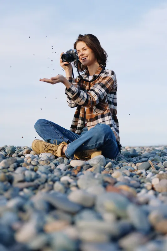 Woman photographer on a rocky beach, capturing moments with a camera as sand and pebbles fly, casual plaid shirt, jeans, and boots under a clear sky.