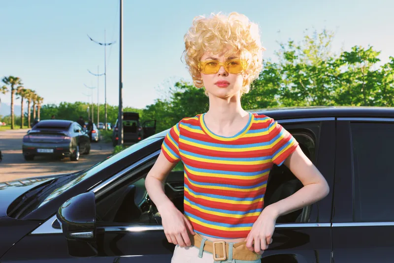 Woman lifestyle scene outdoors with retro vibe, bright striped shirt, curly blonde hair, sunglasses, leaning on a dark car, sunlit moment, relaxed posture, vivid colors, cheerful mood.