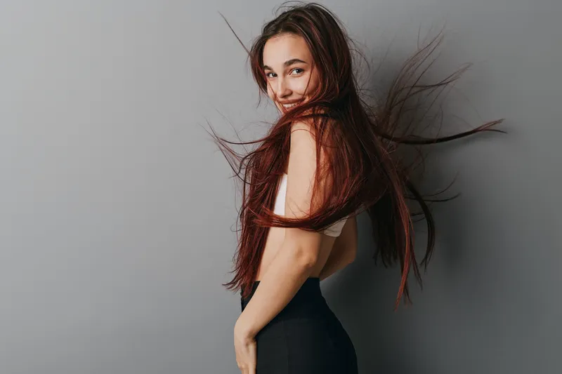 Young woman with long hair posing against gray wall, smiling and looking at camera. Casual outfit with white top and black pants, natural beauty and confident expression.